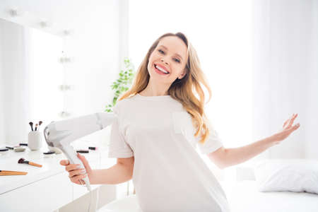 Portrait of attractive dreamy carefree cheerful blond girl drying hair having fun rest at light white home indoorsの写真素材