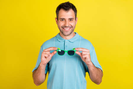 Photo of funny brunet man hold eyewear wear blue t-shirt isolated on yellow color backgroundの写真素材