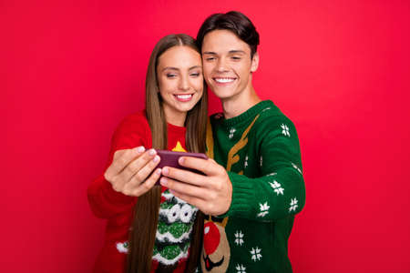 Photo of cheerful nice couple guy hold phone make selfie wear ugly pullover isolated red color backgroundの写真素材