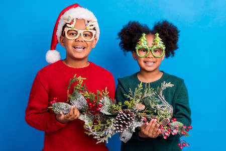 Photo of excited sweet dark skin children wear pullover headwear glasses smiling holding decoration...