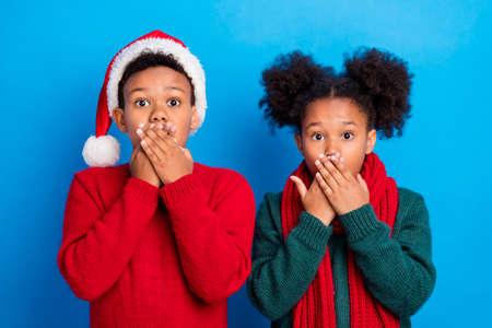 Photo of scared impressed dark skin kids dressed sweaters arms close mouth big eyes isolated blue color backgroundの写真素材