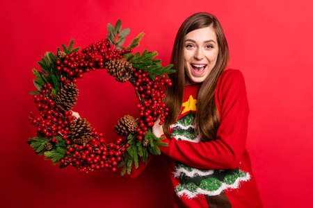 Photo of positive young cheerful woman hold hand wreath smile tradition christmas isolated on red color backgroundの写真素材