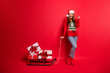 Full body photo of hooray young lady with sledge wear sweater cap jeans boots isolated on red color backgroundの写真素材