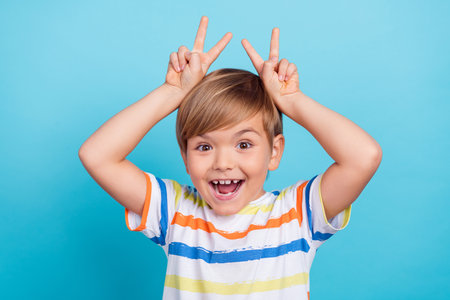 Photo of funky funny schoolboy make v-sign rabbit ear open mouth wear striped t-shirt isolated blue color backgroundの写真素材