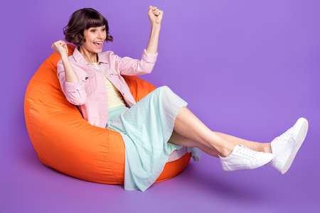 Full size photo of happy cheerful young woman look empty space sit bean bag winner isolated on purple color backgroundの写真素材