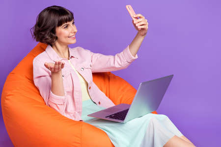 Photo of young woman winner hold hand laptop talk camera phone sit bean bag isolated on violet color backgroundの写真素材