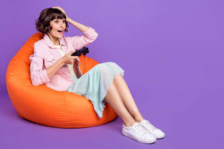 Full body photo of young amazed shocked woman sit bean bag play joystick gamer isolated on purple...