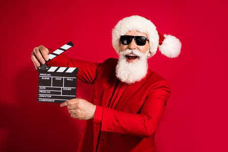 Photo of funny grey hair elder man hold clap wear santa cap red suit eyewear isolated on red color backgroundの写真素材
