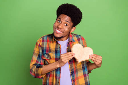 Photo of charming cute dark skin man wear plaid shirt smiling holding wooden heart isolated green...