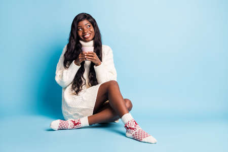 Photo of adorable dark skin lady wear white winter outfit sitting floor coffee looking empty space isolated blue color backgroundの写真素材