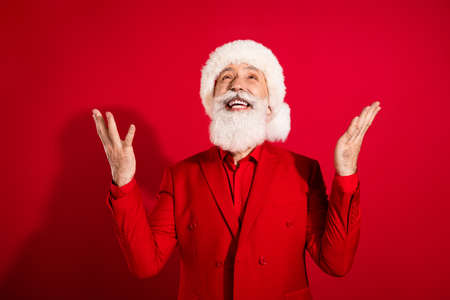 Photo of positive santa claus look up empty space wait raise hands make wish wear x-mas hat suit on red color backgroundの写真素材