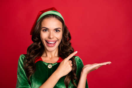 Photo of sweet excited young woman dressed elf costume smiling holding arm pointing empty space isolated red color backgroundの写真素材