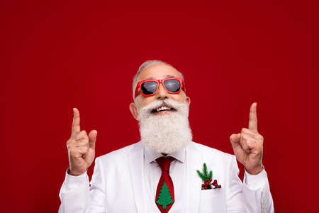 Photo of confident funny man dressed white christmas costume glasses pointing fingers up empty space isolated red color backgroundの写真素材