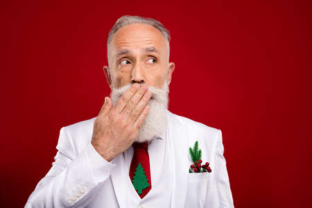 Photo of worried cute mature man dressed christmas costume arm cover lips isolated red color backgroundの写真素材