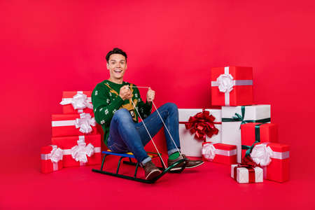 Full length body size photo happy guy in ugly sweater smiling sitting on sledges isolated bright red...