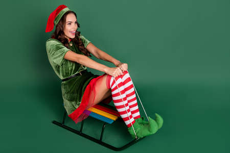 Full length profile side photo of young girl elf happy positive smile have fun ride sledge winter isolated over green color backgroundの写真素材