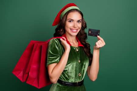 Portrait of beautiful trendy cheerful girl elf holding bank card carrying buyings isolated over green color backgroundの写真素材