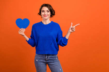 Photo of nice brunette hair millennial lady hold heart point empty space wear blue sweater isolated on orange color backgroundの写真素材