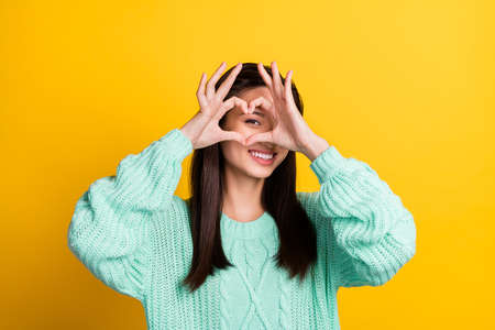 Photo of adorable playful young lady wear turquoise sweater showing fingers heart cover face smiling isolated yellow color backgroundの写真素材