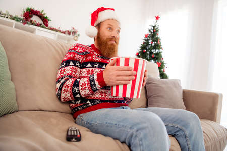 Photo of smiling good mood focused male wear santa claus hat eat food popcorn snack watch movie home cinemaの写真素材