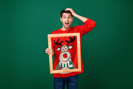 Photo of young excited man hold wooden frame picture wear designed sweater new year isolated over green color backgroundの写真素材