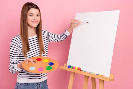 Portrait of attractive cheerful talented girl making new painting picture isolated over pink pastel color backgroundの写真素材
