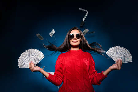 Photo of cool adorable young woman dressed red pullover dark eyewear holding money fans isolated dark blue color backgroundの写真素材