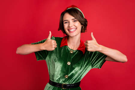 Photo of cheerful young charming lady santa helper make fingers thumbs ups ad isolated on red color backgroundの写真素材