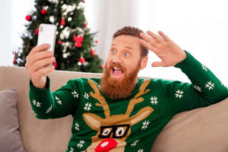 Photo of happy cheerful good mood man take selfie on smartphone showing hand hello greetings relaxing at home houseの写真素材