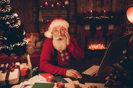 Photo of amazed speechless santa claus take pc delivery order wear hat sweater in north pole office indoorsの写真素材