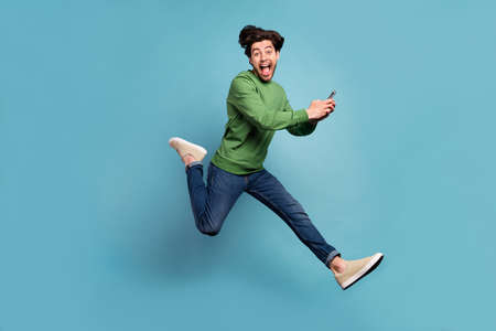 Full size profile side photo of funky excited man jump addicted to social media hold phone isolated on blue color backgroundの写真素材