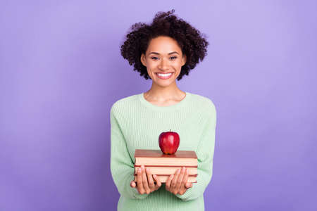 Photo of young cheerful happy afro american woman hold hands books pile apple isolated on purple...