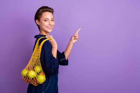 Profile photo of nice brunette millennial lady point empty space hold fruits wear jeans shirt isolated on violet color backgroundの写真素材