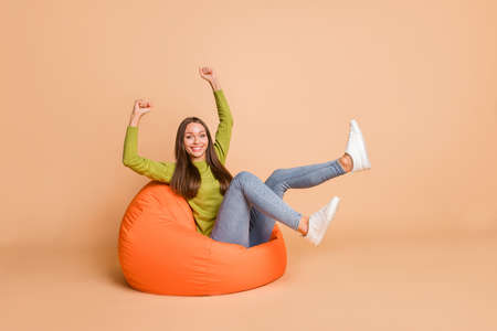 Portrait of nice lovely cheerful girl sitting on orange bag chair celebrating having fun isolated over beige pastel color backgroundの写真素材