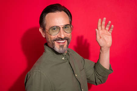 Photo of optimistic age brunet man wave wear khaki shirt eyewear isolated on red color backgroundの写真素材