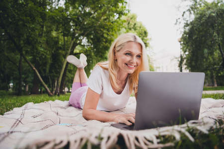 Full body photo of aged nice blond lady sit write laptop wear t-shirt trousers sneakers in park aloneの写真素材