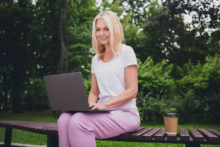 Photo of aged optimistic blond lady sit work laptop wear white t-shirt trousers sit in parkの写真素材
