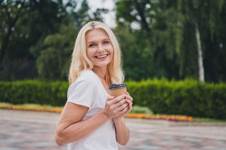 Photo of mature funky blond lady drink coffee wear white t-shirt walk in parkの写真素材