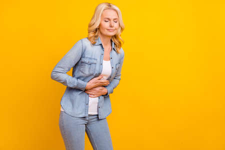 Photo of unhappy sad upset old woman pain bad mood stomach hands isolated on yellow color backgroundの写真素材