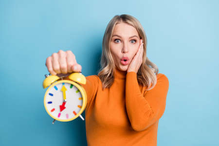 Photo of adorable shocked young woman dressed orange pullover arm cheek showing timer isolated blue color backgroundの写真素材