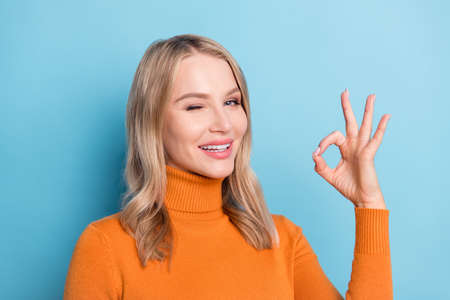 Photo of sweet cute young lady wear orange sweater winking showing okey smiling isolated blue color backgroundの写真素材