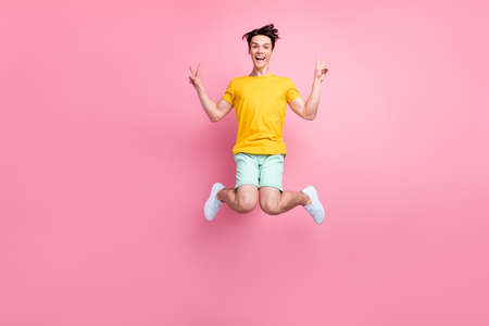 Photo of friendly carefree guy jump show v-signs wear yellow t-shirt shorts footwear isolated on pink backgroundの写真素材