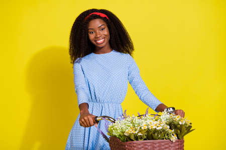 Photo portrait of curly girl wearing headband dotted blue dress riding bike with flowers basket isolated vivid yellow color backgroundの写真素材