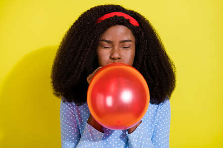 Photo of funky young dark skin positive woman blow air balloon festive decoration isolated on yellow color backgroundの写真素材
