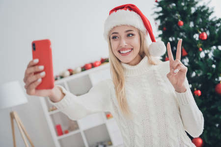 Photo of charming pretty young woman dressed white sweater recording video modern gadget showing v-sign indoors room home houseの写真素材