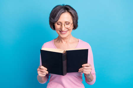 Photo of mature smiling positive dreamy woman in glasses reading interesting book isolated on blue color backgroundの写真素材