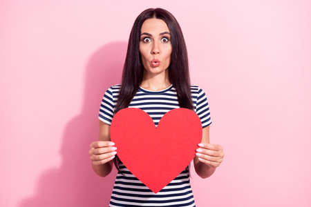 Photo portrait of young girl in striped t-shirt surprised keeping heart card on date isolated on pastel pink color backgroundの写真素材