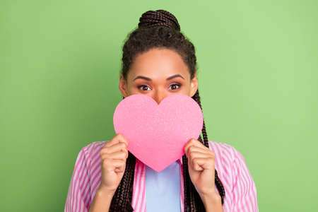 Photo of shy adorable dark skin lady wear pink shirt smiling holding heart cover lips face isolated...