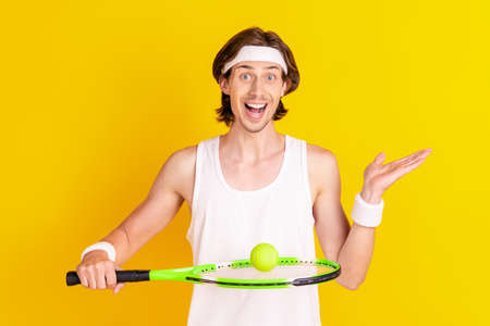 Portrait of attractive skilled cheerful guy playing tennis holding ball on racket isolated over bright yellow color backgroundの写真素材