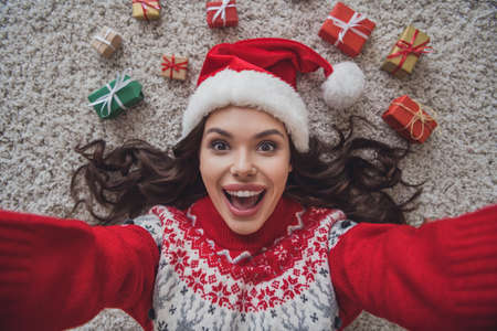 Top view photo portrait smiling amazed woman wearing headwear laying on floor in xmas clothes taking selfieの写真素材